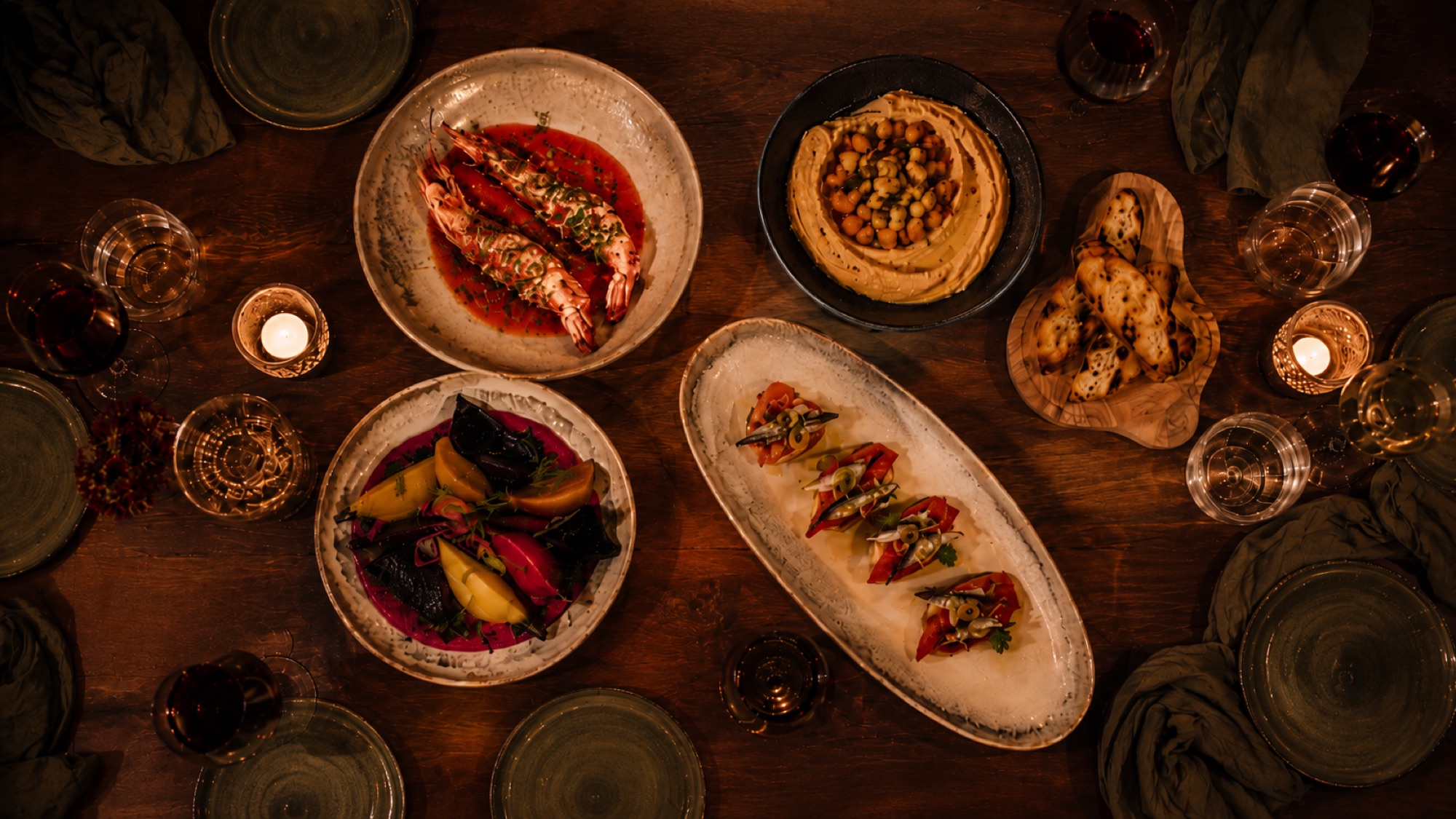 Overhead view of a Mediterranean spread: grilled prawns, hummus, beet salad, and toasts across a warm wooden table